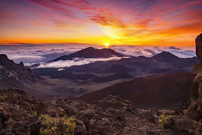 A fiery sunrise glows above a sea of clouds and the cinder-strewn crater at the summit of Haleakalā.