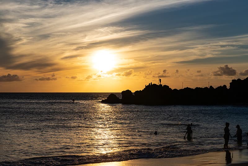 People on the beach watch the sun set beside Black Rock at Kāʻanapali.