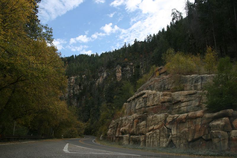 State Route 89A curving past layered sandstone cliffs and autumn trees in Oak Creek Canyon.
